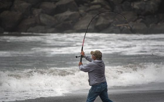 A man in a shirt and hat who is on the seashore and has a fishing rod in his hand.