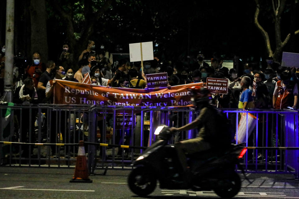 A scooter is driven in the evening in front of people standing behind barricades holding signs that read: Republic of Taiwan welcomes US House Speaker Nancy Pelosi and Madam speaker welcome to Taiwan.